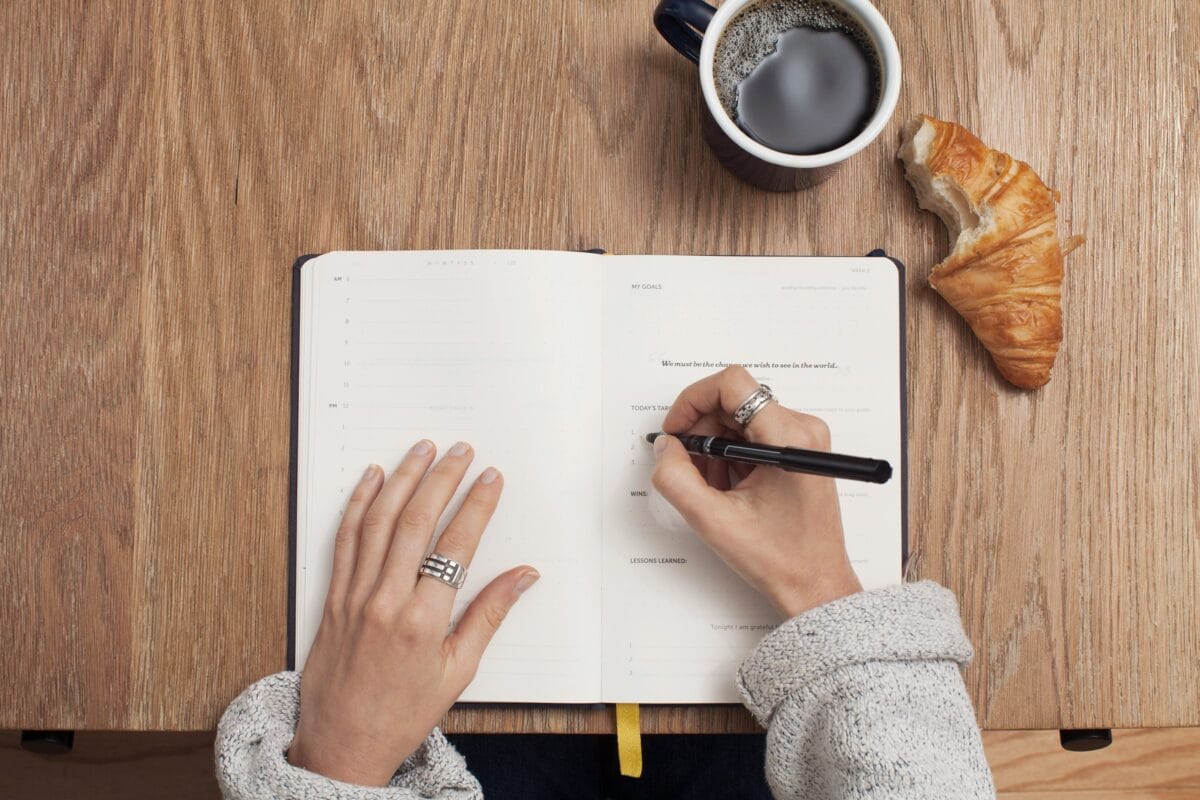 Fotografi av en skrivbok, händer och en penna. Kaffekopp och croissant vid sidan | NTI-skolan