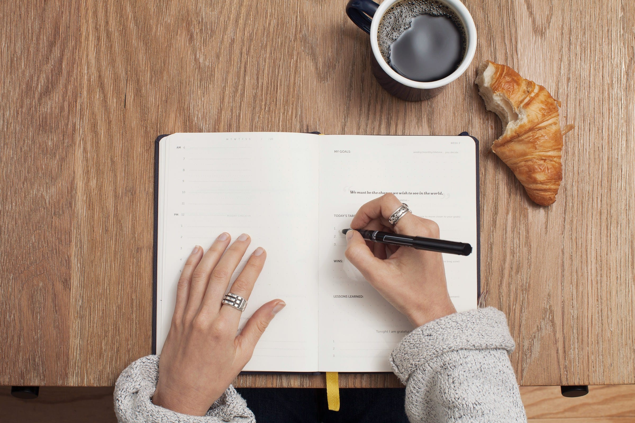 Fotografi av en skrivbok, händer och en penna. Kaffekopp och croissant vid sidan | NTI-skolan