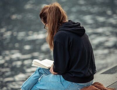En tjej med långt, uppsatt hår sitter utomhus och läser en bok | NTI-skolan